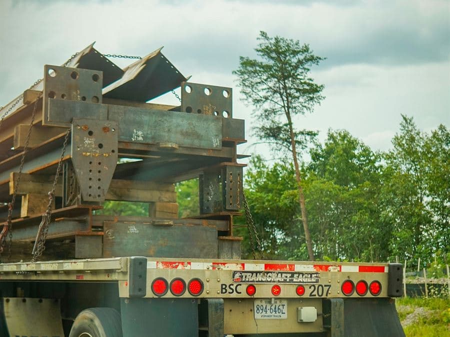 A flatbed trailer loaded with structural steel, seen from the escort vehicle behind it